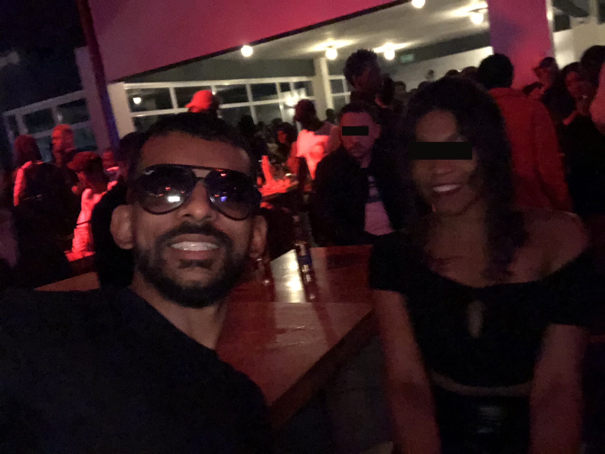Gin in sunglasses next to a woman in a bar, her eyes covered